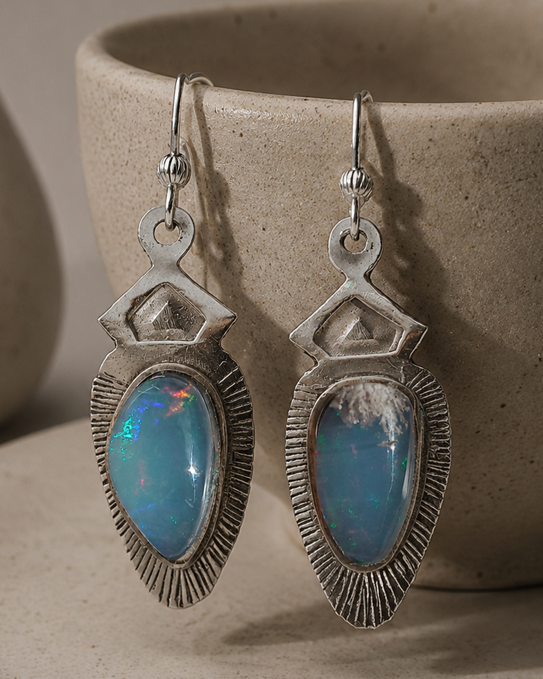 A pair of silver drop earrings with teardrop-shaped blue opal gemstones, displayed on a textured beige ceramic cup resting on a matching saucer.