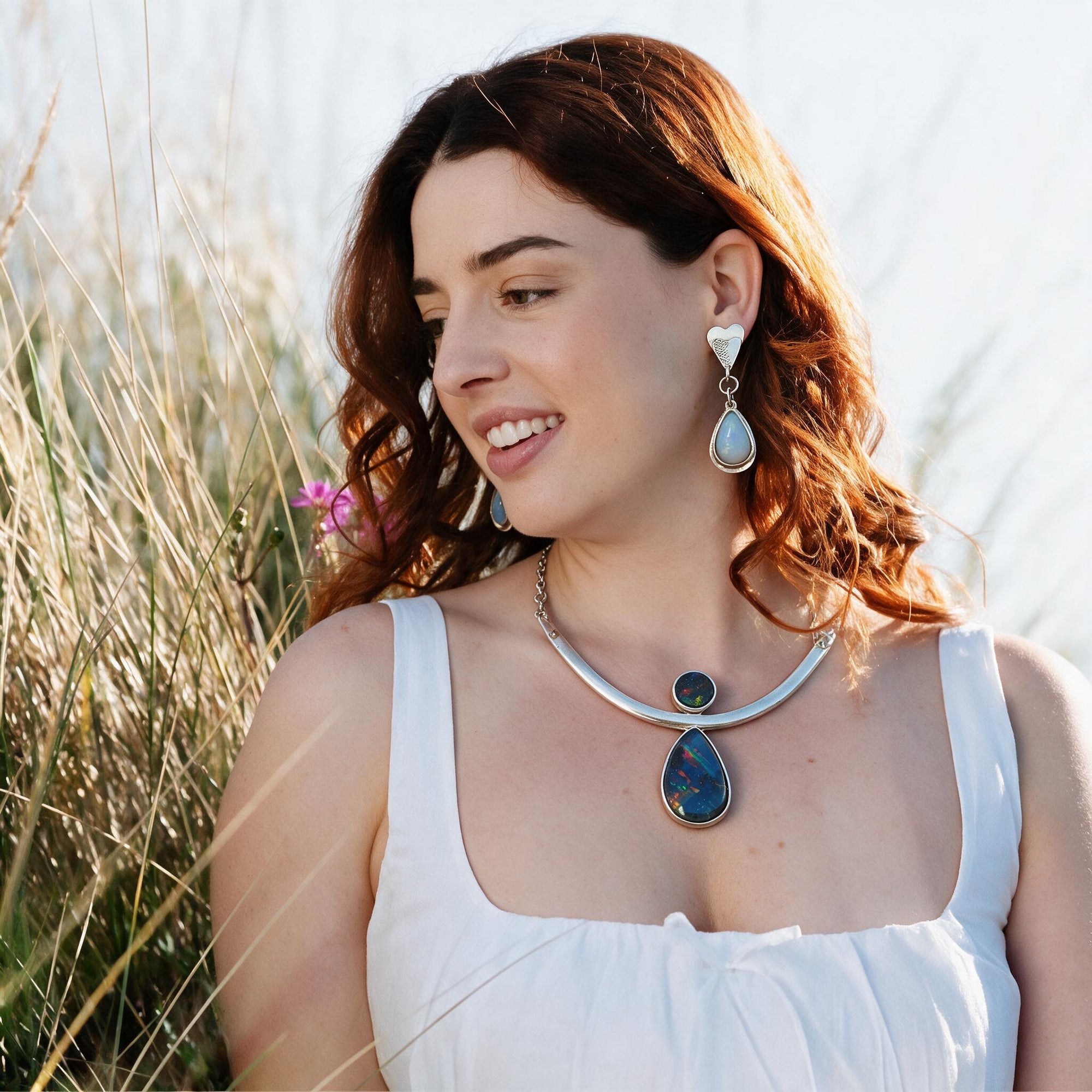 A woman outdoors wearing a silver-toned necklace with a large teardrop-shaped dark blue stone pendant, matching drop earrings, and a white sleeveless top, with dry grass in the background.