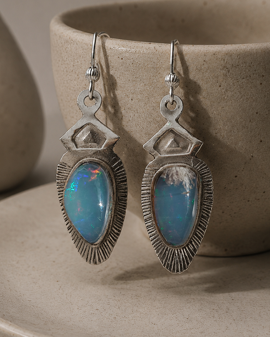 A pair of silver drop earrings with teardrop-shaped blue opal gemstones, displayed on a textured beige ceramic cup resting on a matching saucer.